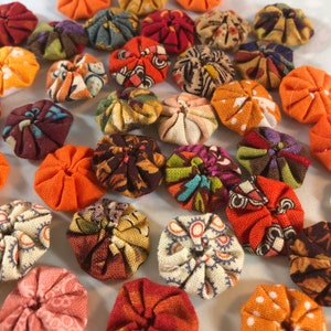 May include: A collection of fabric yo-yos in various colors and patterns. The yo-yos are primarily orange, red, and brown, with some featuring floral and geometric designs. These handmade fabric circles are arranged closely together.