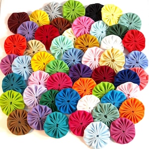 May include: A collection of numerous fabric yo-yos in a wide variety of solid colors including red, blue, yellow, green, pink, purple, orange, brown, black, white, and grey. Each circular piece features gathered fabric radiating from a center point.