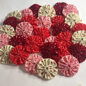 May include: A collection of fabric yo-yos in shades of red and cream. Some yo-yos feature a heart pattern, while others are solid red. These handmade fabric circles are ideal for crafting and sewing projects.
