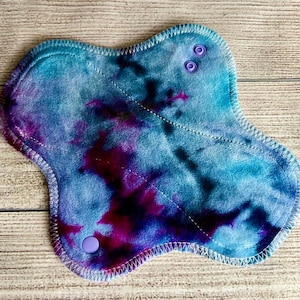 May include: A reusable cloth menstrual pad featuring a tie-dye pattern in shades of blue, purple, and magenta. The pad has a contoured shape with stitched edges and two purple plastic snap fasteners located at the top and bottom wings.