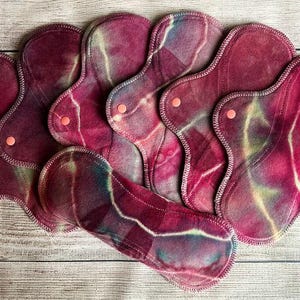 May include: A collection of seven reusable cloth menstrual pads featuring a tie-dye pattern in shades of deep magenta, burgundy, and hints of teal. Each pad has a contoured shape with stitched edges and a small, round, light orange plastic snap fastener.