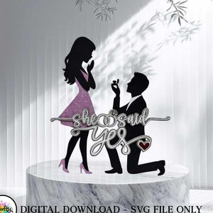 May include: A black and silver cake topper with the words "she said yes" and silhouettes of a kneeling man and a woman in a pink dress. A red heart is included in the design. The cake topper is on a white marble stand.