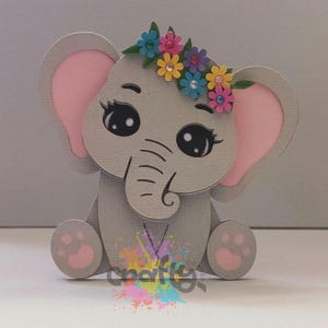 May include: A gray elephant-shaped papercraft with pink inner ears and feet. The elephant has large, expressive eyes and a trunk. A colorful floral crown adorns its head. The design is whimsical and suitable for children's decor.