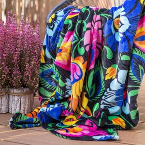 Può includere: Una coperta colorata con motivi floreali su sfondo nero. La coperta presenta grandi fiori nei toni del blu, rosa, giallo e arancione, con foglie verdi. La coperta è drappeggiata su una sedia e poggia su una superficie di legno.