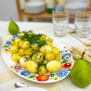 Può includere: Un piatto ovale bianco con un motivo floreale colorato contiene patate bollite condite con aneto fresco. Due pere verdi e due bicchieri vuoti sono sullo sfondo. Il piatto è il punto focale dell'immagine.