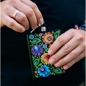 Puede incluir: Petaca negra con un diseño floral colorido con flores azules, moradas, amarillas y naranjas. La petaca se está abriendo, revelando una tapa plateada. El patrón floral se encuentra sobre un fondo negro.