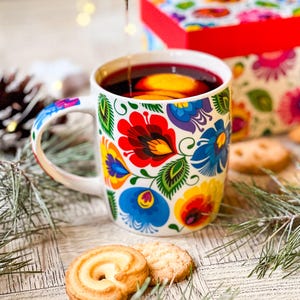 Könnte beinhalten: Ein weißer Keramikbecher mit einem lebendigen, mehrfarbigen Blumenmuster im Volkskunststil in Rot, Blau, Gelb und Grün. Der Becher ist mit einer dunkelroten Flüssigkeit gefüllt, in der Orangenscheiben schwimmen, und hat einen passenden Henkel.
