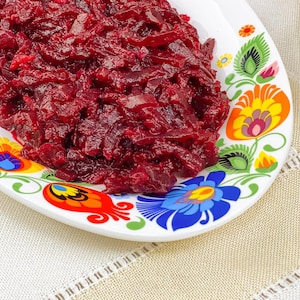 Può includere: Un piatto di vivace insalata di barbabietole rosse. L'insalata è finemente sminuzzata e appare lucida. Il piatto bianco è decorato con motivi floreali colorati in giallo, blu, verde e arancione. Il piatto è su una tovaglia beige.