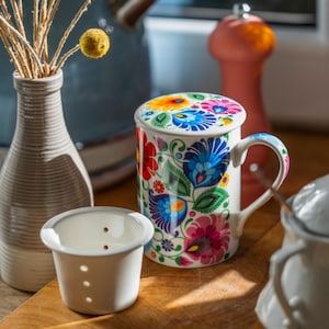 Op de afbeelding: Een witte keramische mok met een bloemmotief in levendige kleuren, waaronder blauw, roze en geel, wordt getoond met een bijpassende deksel. Een kleine witte thee-infuser en een vaas met gedroogde bloemen zijn ook zichtbaar.