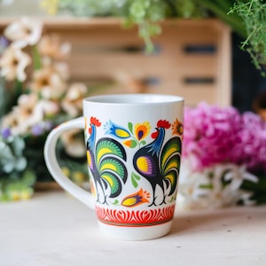 Può includere: Tazza in ceramica bianca con un colorato motivo a gallo con accenti rossi, blu, gialli e verdi. La tazza ha un manico curvo e una fascia a motivi rossi e verdi alla base. Fiori e una cassa di legno sullo sfondo.