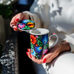 Può includere: Una tazza in ceramica con coperchio, caratterizzata da un vivace motivo floreale in rosso, blu, arancione e rosa su sfondo nero. La tazza ha interno e manico bianchi. Il coperchio riprende il motivo floreale. Un oggetto decorativo e colorato.