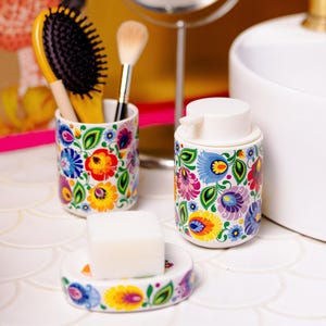 Può includere: Un set da bagno in ceramica con un colorato motivo floreale. Il set comprende un portaspazzolino con spazzola per capelli e pennello per il trucco, un dosatore di sapone e un portasapone con una saponetta bianca. Il motivo floreale comprende fiori blu, gialli e viola.