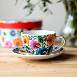 Può includere: Un set di tazza e piattino bianchi con un vivace motivo floreale nei toni del blu, arancione, giallo e rosa. La tazza ha un manico curvo e poggia su un piattino abbinato. Una scatola decorativa con bordo rosso è sullo sfondo.