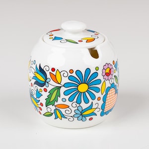 Op de afbeelding: Een witte keramische suikerpot met een ronde vorm en een bijpassend deksel. Het oppervlak is voorzien van een levendig, veelkleurig bloemenpatroon met blauwe, gele, oranje, groene en roze bloemblaadjes, bladeren en stippen op een witte achtergrond.
