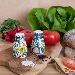 May include: Two white ceramic salt and pepper shakers featuring a colorful hand-painted floral and heart pattern in shades of blue, yellow, green, and orange. The shakers have a rounded, tapered shape and are positioned on a wooden cutting board.