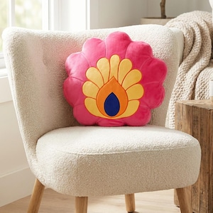Può includere: Un cuscino decorativo a forma di fiore rosa con dettagli gialli, arancioni e blu è appoggiato su una sedia color crema. Il design del cuscino ricorda un fiore stilizzato, aggiungendo un tocco di colore all'ambiente neutro. La sedia ha gambe in legno.