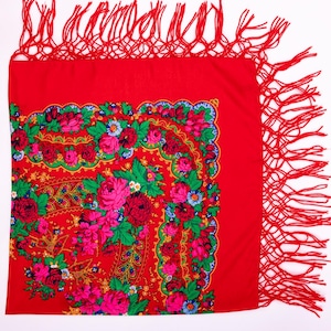Op de afbeelding: Een vierkante rode sjaal met een levendig bloemenpatroon van grote roze, rode en donkerrode rozen, groene bladeren en kleine blauwe en witte bloemetjes. De sjaal is afgewerkt met lange, dunne rode franjes langs twee aangrenzende zijden.
