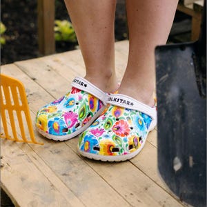 Peut inclure: Une paire de sabots blancs à enfiler arborant un motif floral folklorique multicolore et vif sur toute la surface. Chaque chaussure possède une bride de talon blanche avec le texte FOLKSTAR et une étoile imprimés en noir sur le côté extérieur.