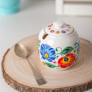 Peut inclure: Un sucrier en céramique blanche de forme arrondie avec un couvercle assorti, orné de motifs floraux aux couleurs vives bleu, rouge, vert et jaune. Une petite encoche sur le bord permet d'accueillir une cuillère en métal argenté au manche orné.