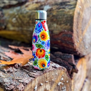 Può includere: Una borraccia in acciaio inossidabile con tappo argentato, ornata da un vivace motivo floreale nei toni del rosso, giallo, blu e viola. La borraccia è appoggiata su tronchi di legno, mettendo in risalto il suo design colorato.