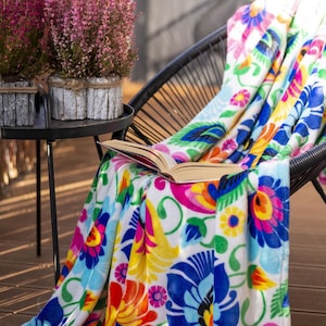 Può includere: Una coperta colorata a motivi floreali drappeggiata su una sedia in vimini nera. La coperta presenta uno sfondo bianco con vibranti fiori blu, rosa, gialli e verdi. Un libro aperto è appoggiato sulla coperta. Due piante in vaso con fiori rosa sono su un tavolino nero.
