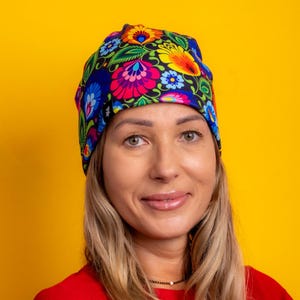 Op de afbeelding: Een kleurrijke beanie-muts met een bloemenpatroon in roze, blauw, geel en groen. De muts staat tegen een heldergele achtergrond. De vrouw op de foto draagt een rood shirt.