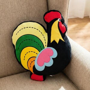 Può includere: Un cuscino a forma di gallo in peluche con corpo nero, cresta rossa e becco giallo. Le piume della coda sono un vivace mix di giallo, verde e arancione, con accenti blu e rosa. Il cuscino è decorativo.