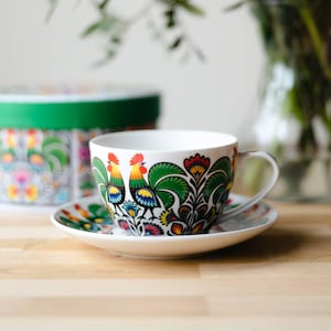 Può includere: Servizio da tè in ceramica bianca con un motivo colorato di gallo e fiori. La tazza e il piattino presentano un vivace motivo di galli, fiori e foglie in rosso, verde, giallo e blu. Una scatola rotonda abbinata è visibile sullo sfondo.