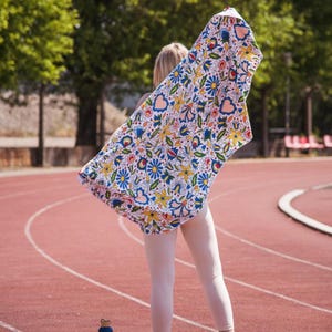 Könnte beinhalten: Ein buntes Handtuch mit Blumenmuster und blauen, gelben und roten Blumen. Das Handtuch wird in die Luft gehalten, wodurch das Muster zur Geltung kommt. Ein helles Accessoire für Strand oder Pool, mit weißem Hintergrund.