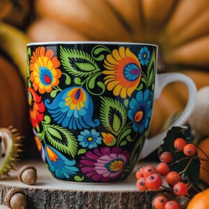 Könnte beinhalten: Eine schwarze Keramik-Kaffeetasse mit einem lebendigen Blumenmuster in Blau-, Orange-, Gelb- und Lilatönen. Die Tasse hat einen weißen Henkel und steht vor einem verschwommenen Hintergrund aus Kürbissen und Herbstlaub.