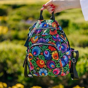 Peut inclure: Un sac à dos coloré avec un motif floral dans les tons de rouge, bleu, jaune et violet sur un fond noir. Le sac à dos comporte plusieurs compartiments zippés, des bretelles noires et une poignée supérieure. Le sac est tenu par une personne.