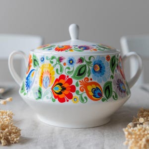 Può includere: Zuccheriera in ceramica bianca con coperchio, ornata da un vivace motivo floreale in rosso, arancione, giallo, blu e verde. La ciotola ha due manici e un pomello sul coperchio. Il design ricorda l'arte popolare.