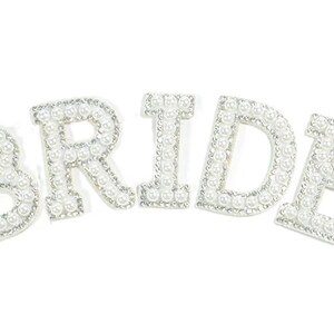 May include: The word BRIDE spelled out in five individual letters. Each letter is constructed with a white pearl centre surrounded by a border of small, clear, reflective rhinestones, creating a decorative and textured appearance for each character.