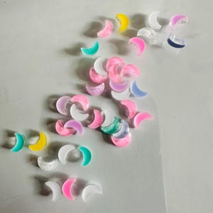 Puede incluir: Una colección de dijes de resina en forma de media luna de colores, en varios tonos de rosa, azul, amarillo y blanco. Los dijes están esparcidos sobre una superficie blanca.
