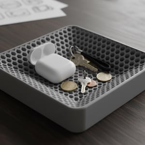 Può includere: Un vassoio quadrato grigio con un motivo a nido d'ape contiene una custodia bianca per auricolari wireless, chiavi e monete. Il vassoio è su una superficie di legno scuro, offrendo un'elegante soluzione di archiviazione.