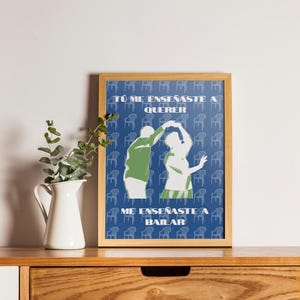 May include: A framed art print featuring a couple dancing, with the text "Tú me enseñaste a querer" and "Me enseñaste a bailar" in white. The background is blue with a pattern of small blue chairs. The frame is a light brown color.
