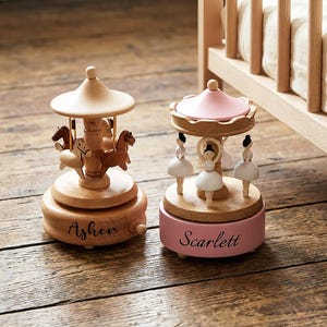 Op de afbeelding: Twee houten carrouselmuziekdozen. Eén heeft paarden en de naam "Asher". De andere heeft ballerinafiguren en de naam "Scarlett". Beide hebben een lichte houten afwerking en staan op een houten oppervlak. Eén carrousel heeft een roze bovenkant.