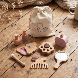 Könnte beinhalten: Ein Holzspielzeug-Beauty-Set mit einem Kordelzugbeutel, Nagellack, Lippenstift, einer Parfümflasche, einem Kamm, einem Spiegel und einem Puder. Das Set ist aus Holz gefertigt und hat eine natürliche Oberfläche. Der Beutel hat eine Zeichnung.