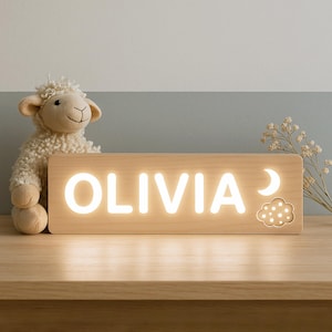 以下が含まれることがあります： 「OLIVIA」という名前が白く光る木製のナイトライト。三日月と雲のデザインが含まれています。左側には羊のぬいぐるみがあります。ライトは木製の表面に置かれています。
