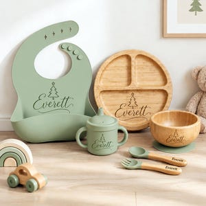 Può includere: Un bavaglino in silicone verde salvia, un piatto diviso in bambù, una ciotola in bambù con base verde, un bicchiere con beccuccio verde salvia e un set di cucchiaio e forchetta in bambù. Ogni oggetto reca il nome Everett e un albero di Natale.