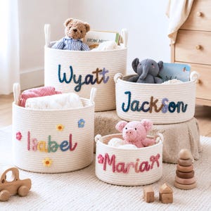 May include: Four round, off-white woven rope storage baskets with handles. Each basket features a personalized name in colorful yarn: Wyatt, Jackson, Isabel, and Maria. The baskets contain plush toys, books, and blankets, with small flower accents.