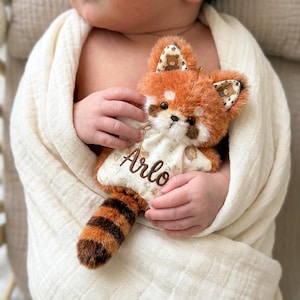 Può includere: Un peluche di panda rosso con pancia bianca e coda a strisce. Il giocattolo ha il nome "Arlo" ricamato sul petto. Il panda rosso è tenuto in una morbida coperta color crema.