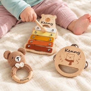 Könnte beinhalten: Ein bärenförmiges Holz-Xylophon mit fünf bunten Metallklangplatten, einem Holzschlägel, einem runden Holz-Tamburin mit Bärenmotiv und einer braunen gehäkelten Bärenrassel. Jeder Gegenstand ist mit dem Namen Kenny in schwarzer Schrift personalisiert.