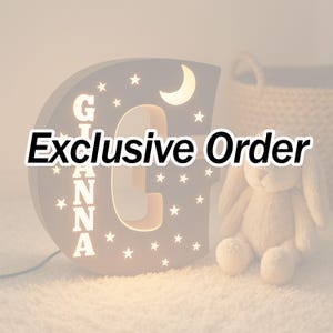 Könnte beinhalten: Nachtlicht in Form eines marineblauen Buchstabens "G" mit dem Namen "GIANNA" in weißen Buchstaben. Das Licht hat Mond- und Sternaussparungen. Der Text "Exclusive Order" wird angezeigt. Ein Plüschhase sitzt in der Nähe.