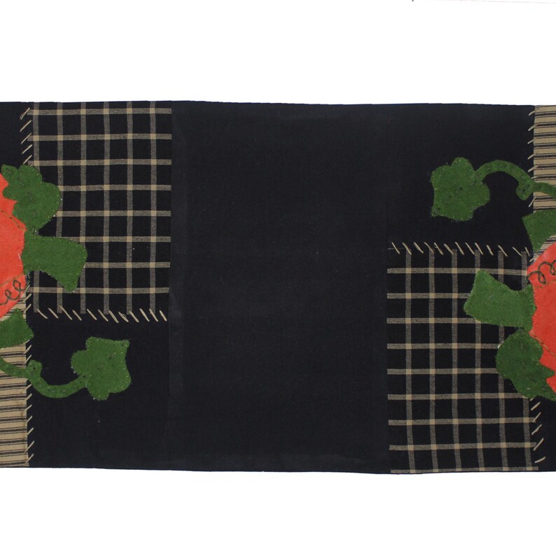 Puede incluir: Un camino de mesa negro con tem&aacute;tica de calabaza. El camino de mesa presenta un gran panel central negro, flanqueado por secciones de tela a cuadros y a rayas. Calabazas naranjas con hojas verdes est&aacute;n aplicadas en los laterales.