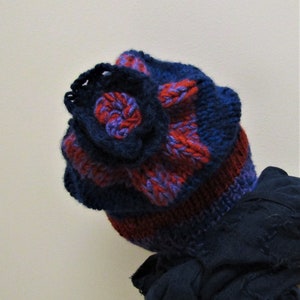 May include: A blue and red striped knitted hat with a blue flower on top. The hat is folded over and the flower is facing the camera.