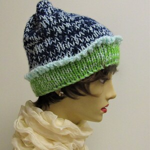 Op de afbeelding: Een blauwe en witte gebreide beanie met een groene en witte gebreide rand. De beanie heeft een lichtblauwe gehaakte rand.