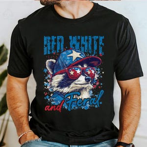 Puede incluir: Camiseta negra con un mapache con gafas de sol y un sombrero con dise&ntilde;o de estrella. El texto de la camiseta dice "Red White and Feral" en azul y rojo. El mapache lleva rojo, blanco y azul.