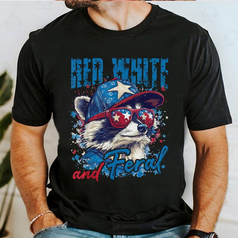 Puede incluir: Camiseta negra con un mapache con gafas de sol y un sombrero con dise&ntilde;o de estrella. El texto de la camiseta dice "Red White and Feral" en azul y rojo. El mapache lleva rojo, blanco y azul.