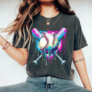 May include: Dark gray t-shirt featuring a baseball design. The graphic includes a baseball with dripping paint, crossed bats, and a shield shape, in shades of blue, pink, and white. The shirt is a casual style.